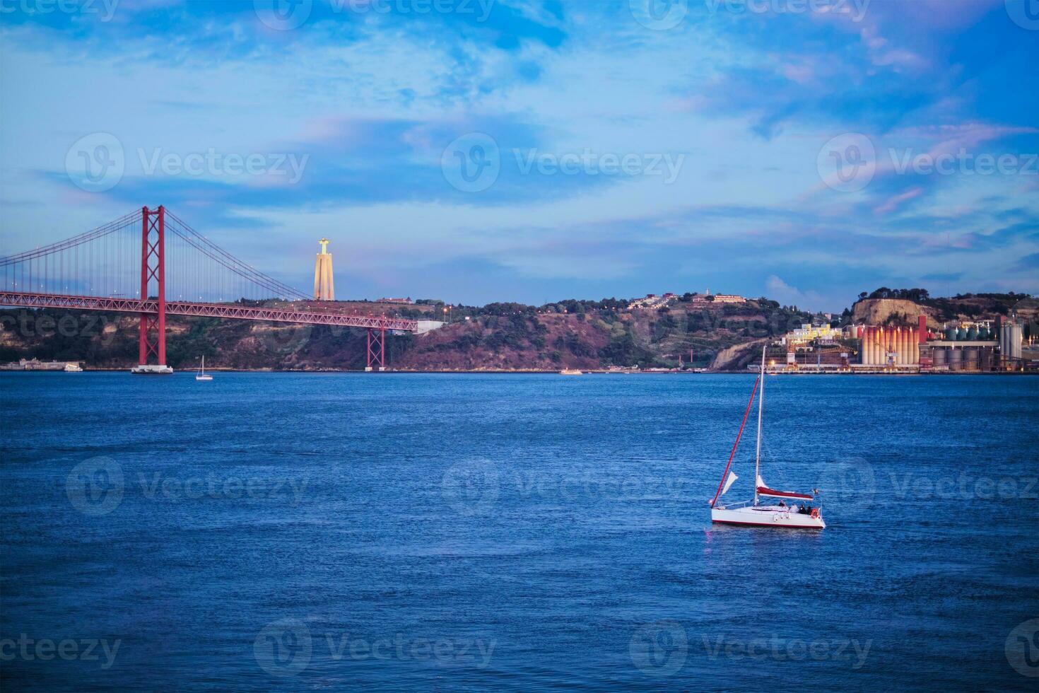 View of 25 de Abril Bridge over Tagus river, Christ the King statue and yacht boat in evening twilight. Lisbon, Portugal photo