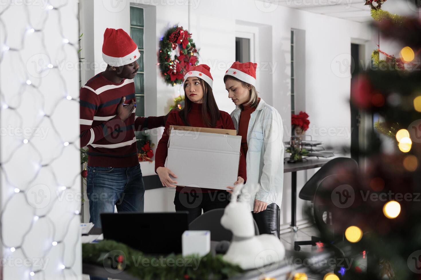 Asian employee getting consoled by colleagues after being fired. Worker holding box filled with desk belongings saying goodbye to coworkers after being laid off from Christmas decorated workplace photo