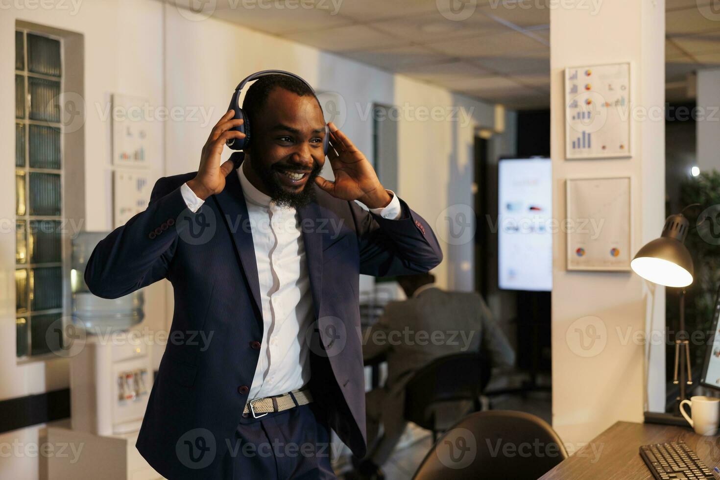 Cheerful manager wearing hadphone listening music in startup office late at night. African american entrepreneur dancing and having fun after finishing financial report. Business concept photo