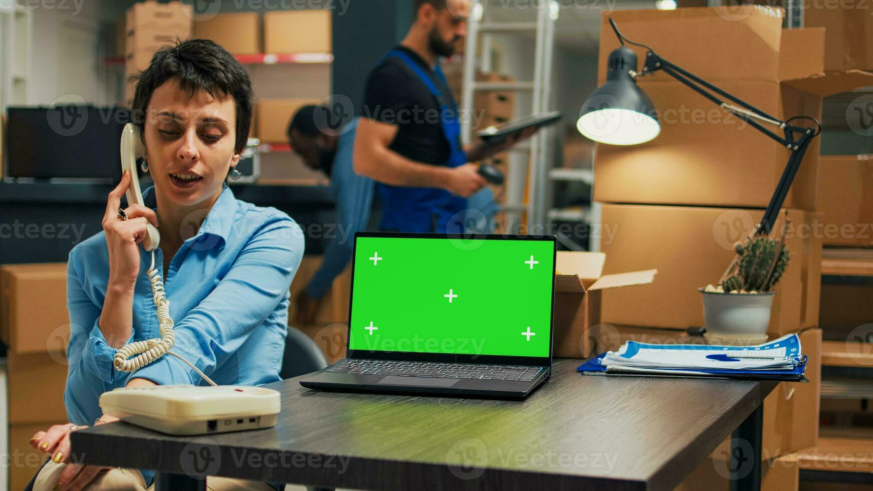 Female entrepreneur answering landline phone in storehouse space and using greenscreen display on laptop. Woman chatting on telephone with cord and looking at isolated mockup screen. photo