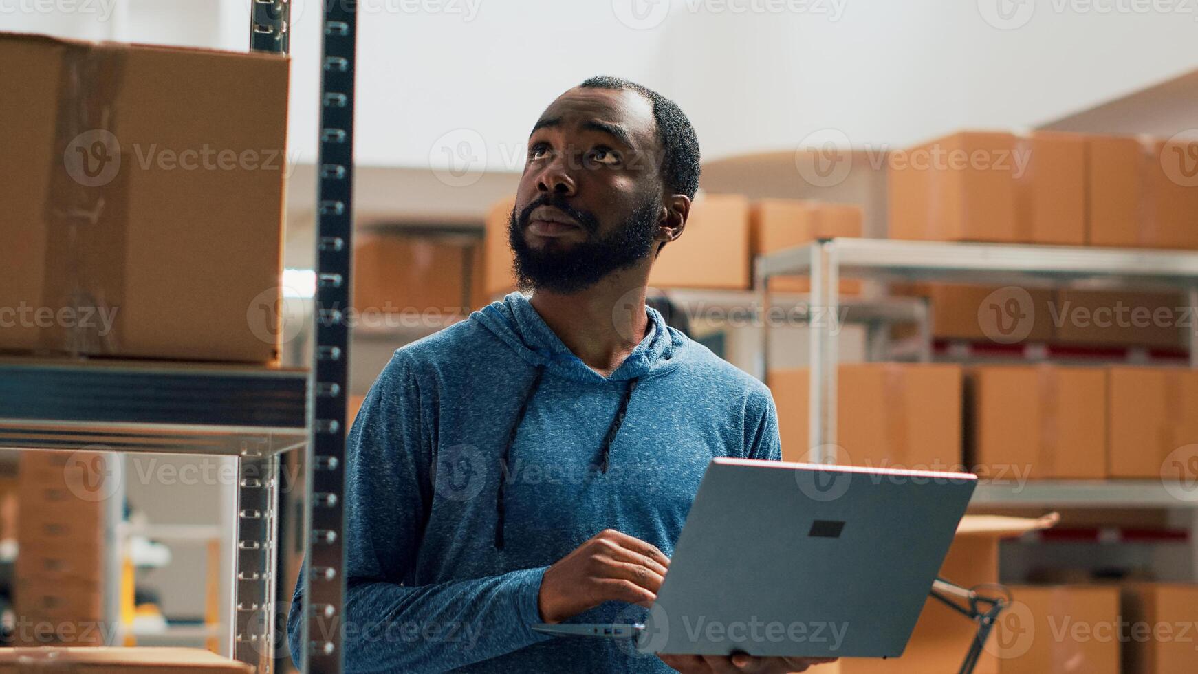 African american man working on merchandise inventory using laptop and taking notes in storehouse. Entrepreneur checking products in cardboard boxes, working on quality control and distribution. photo