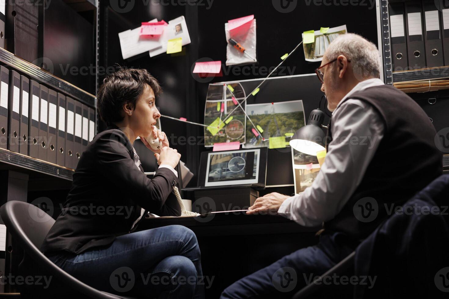 Woman investigator discussing with remote inspector at landline phone, working overtime at criminal case in arhive room. Criminology team analyzing crime scene evidence, checking federal report photo