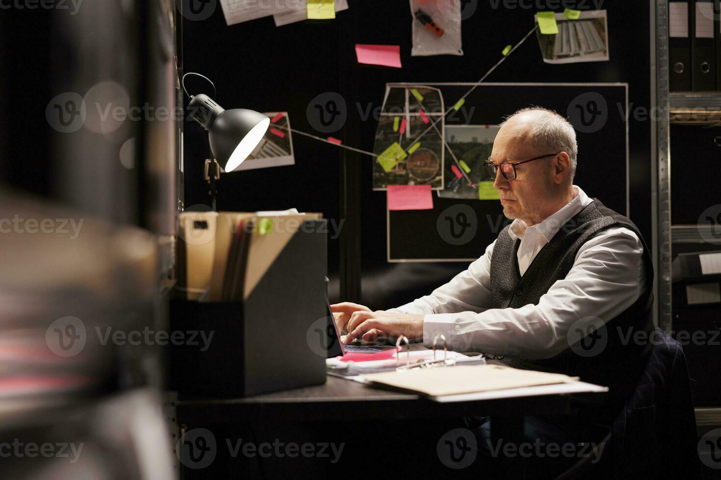 Elderly man inspector sitting at desk table in evidence room, working late at night at missing person case. Senior private detective analyzing victim files, looking at crime scene report photo