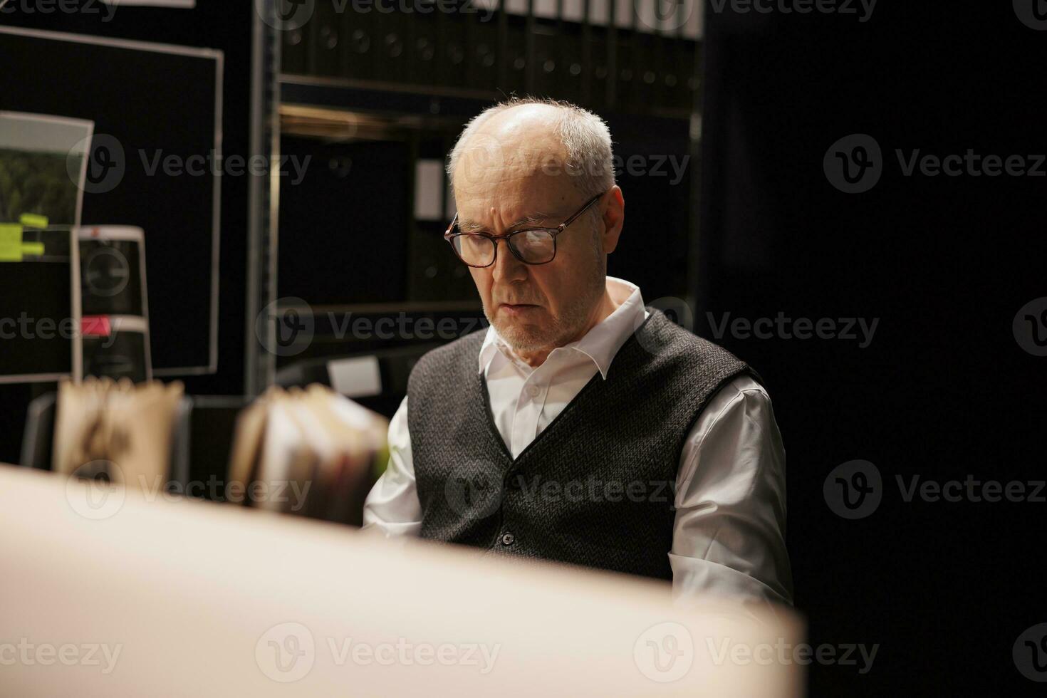 Elderly investigator looking at criminal investigations files, working overtime at mysterious suspect profile in arhive room. Senior police officer analyzing crime scene evidence late at night photo