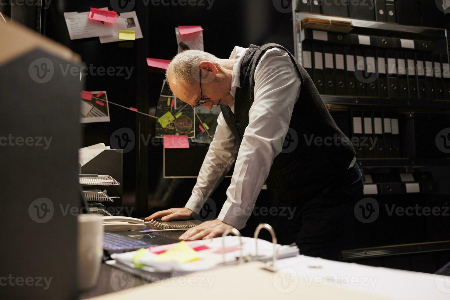 Inspector working overtime at criminal investigations case, analyzing evidence files in arhive room. Senior private detective checking criminology report, standing in police department photo