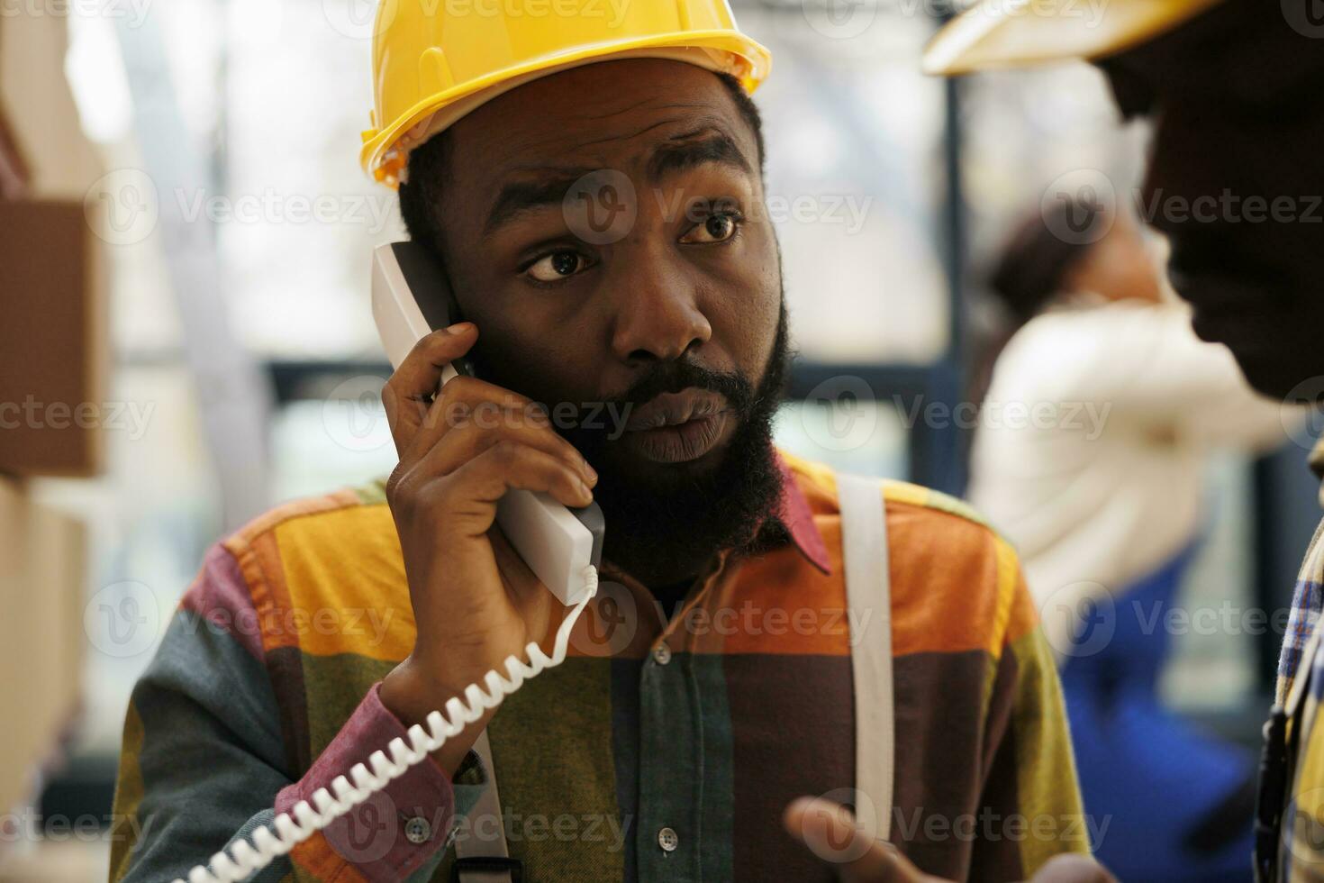 African american man talking on landline phone in industrial storehouse. Factory warehouse operator answering supervisor telephone call and listening to inventory management instructions photo