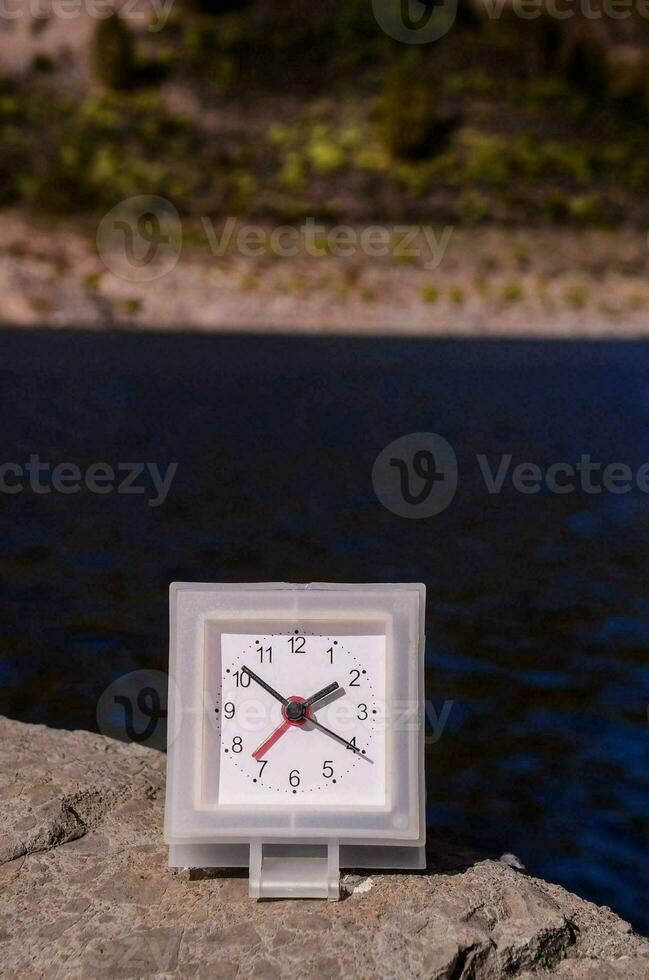 a small clock sitting on a rock near a lake photo