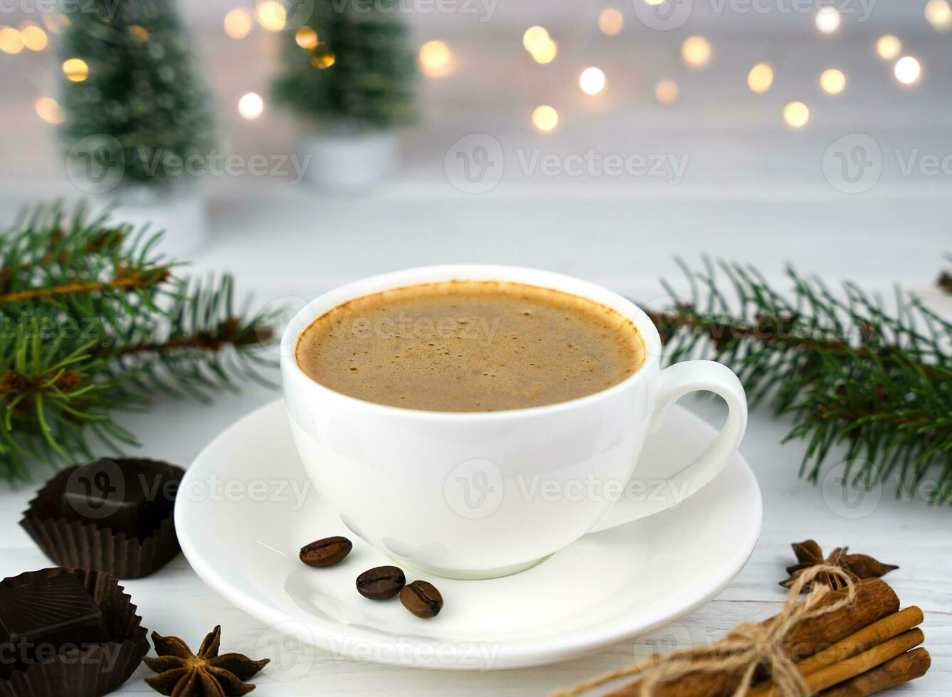 Cup of fresh coffee, chocolate candies and cinnamon on a white wooden table. Christmas lights garland and Christmas decor on background. Close-up. Selective focus. photo