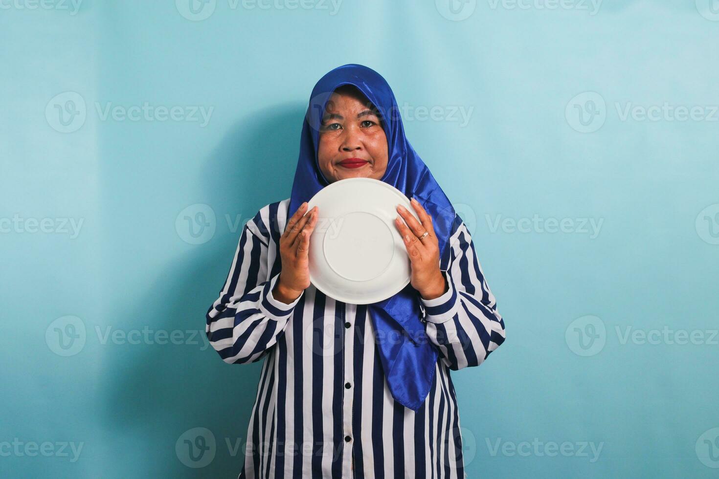 An unhappy middle-aged Asian woman in a blue hijab and a striped shirt is holding an empty white plate, feeling hungry during a diet or fasting. She is isolated on a blue background photo