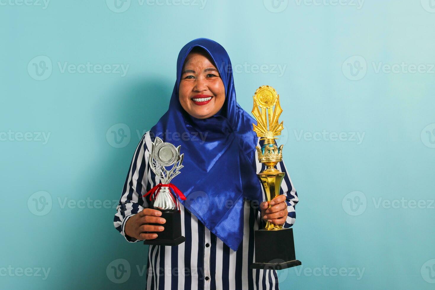 An excited middle-aged Asian businesswoman in a blue hijab and a striped shirt is holding a gold and silver trophy, celebrating her success and achievement. She is isolated on a blue background photo