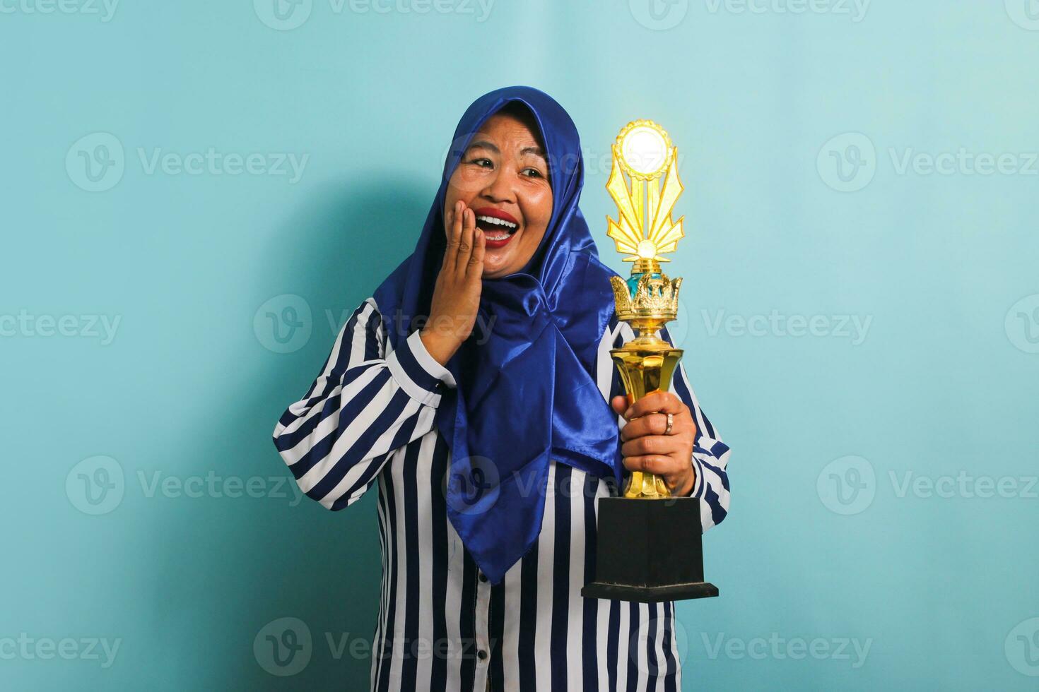 un emocionado de edad mediana asiático mujer de negocios en un azul hijab y un a rayas camisa es participación un oro trofeo, celebrando su éxito y logro. ella es aislado en un azul antecedentes foto