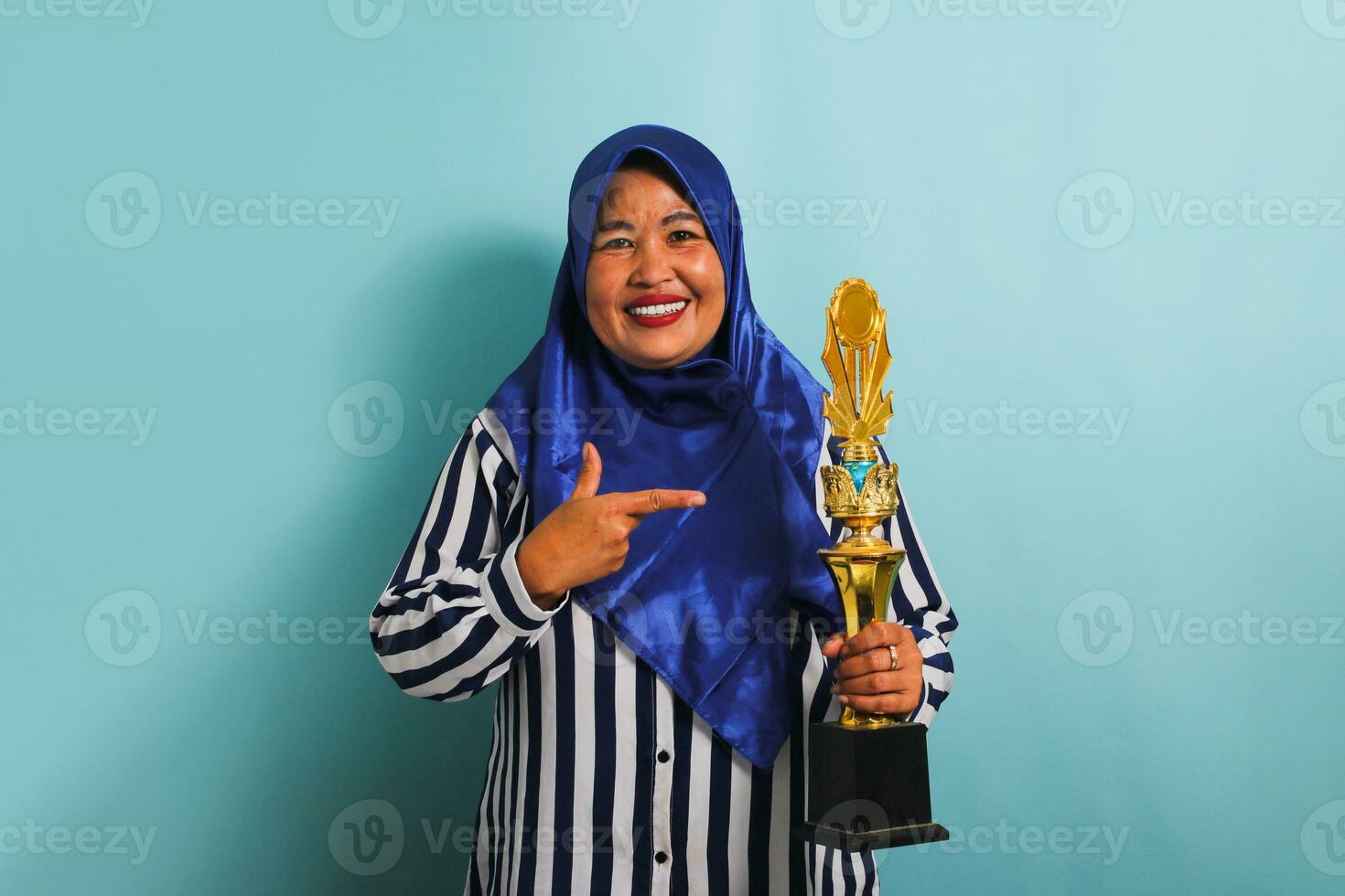 A happy middle-aged Asian businesswoman in a blue hijab and a striped shirt is holding a gold trophy and pointing at it, celebrating her success and achievement. She is isolated on a blue background photo