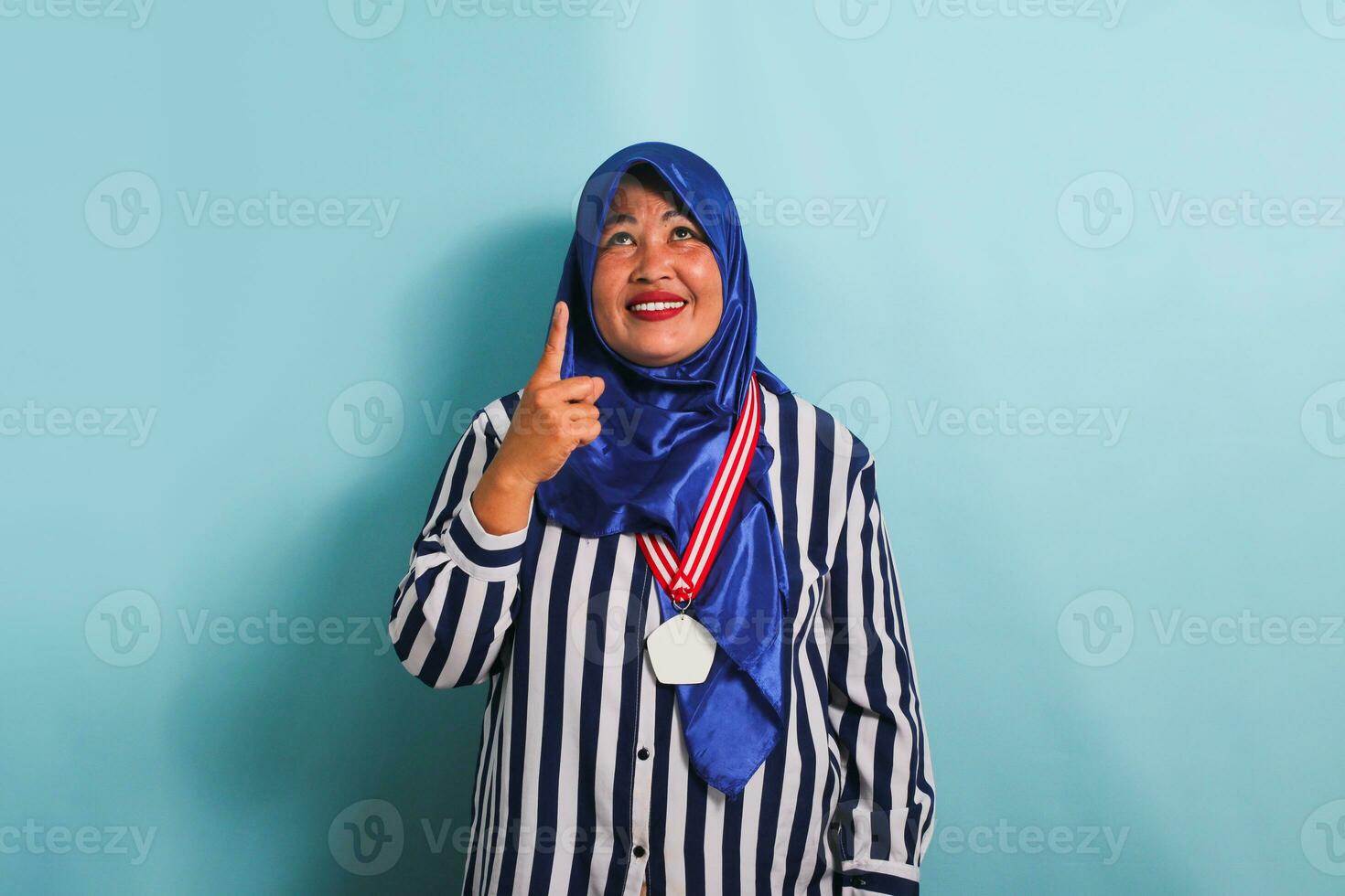 A happy middle-aged Asian woman, wearing a blue hijab and a striped shirt, is pointing her finger upwards towards the copyspace. She is wearing a medal and is isolated on a blue background photo