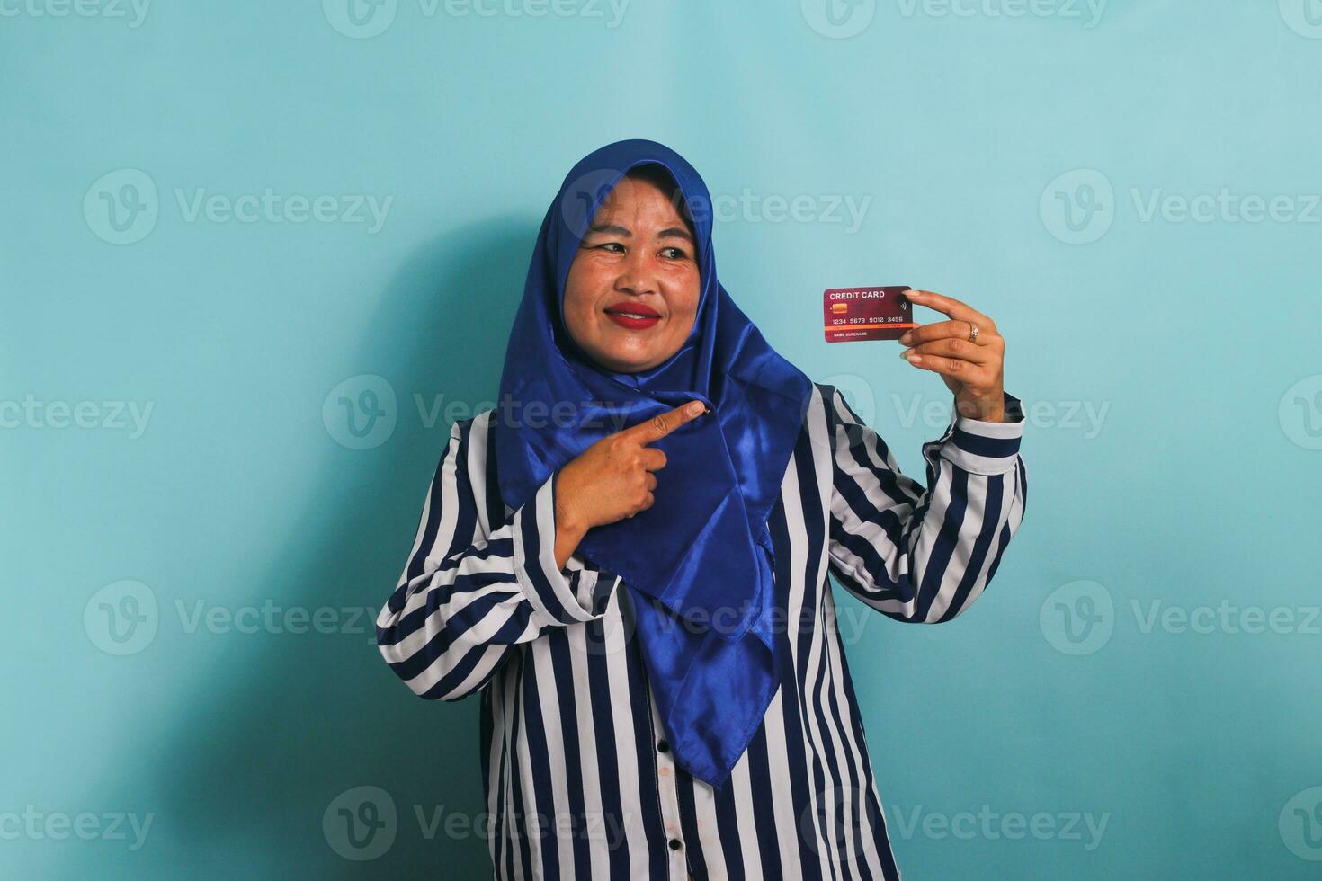 emocionado de edad mediana asiático mujer en azul hijab y a rayas camisa señalando dedo a crédito tarjeta, recomendando pago método, yendo en compras, aislado terminado azul antecedentes. foto