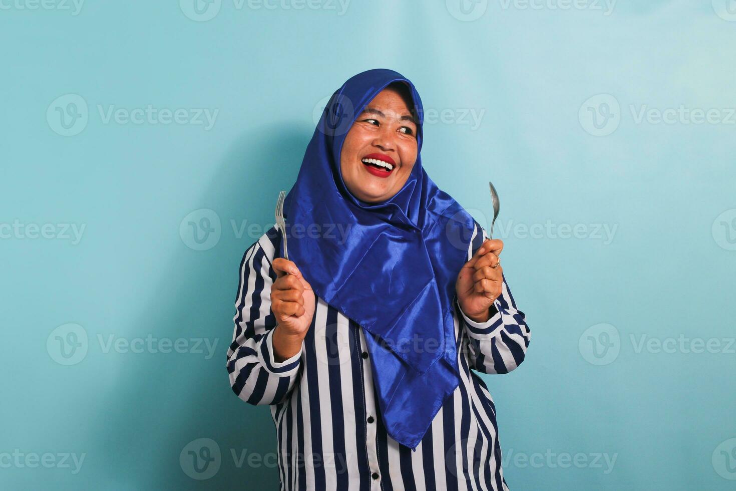 An excited middle-aged Asian woman in a blue hijab and a striped shirt is holding a spoon and fork, feeling hungry while waiting for food to be served. She is isolated on a blue background photo