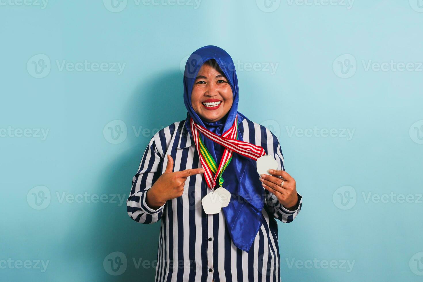 An excited middle-aged Asian woman, wearing a blue hijab and a striped shirt, is proudly pointing to the medal she is holding, isolated on blue background. photo
