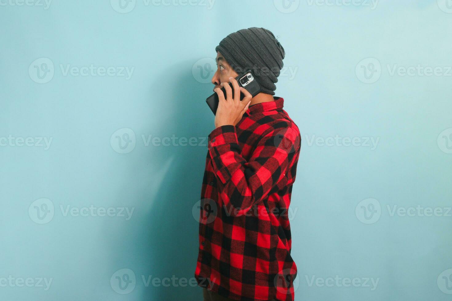 conmocionado joven asiático hombre con gorro sombrero y rojo tartán franela camisa es sorprendido, con su mano en su cintura en un brazo en jarras posición, mientras hablando en su móvil teléfono, aislado en un azul antecedentes foto