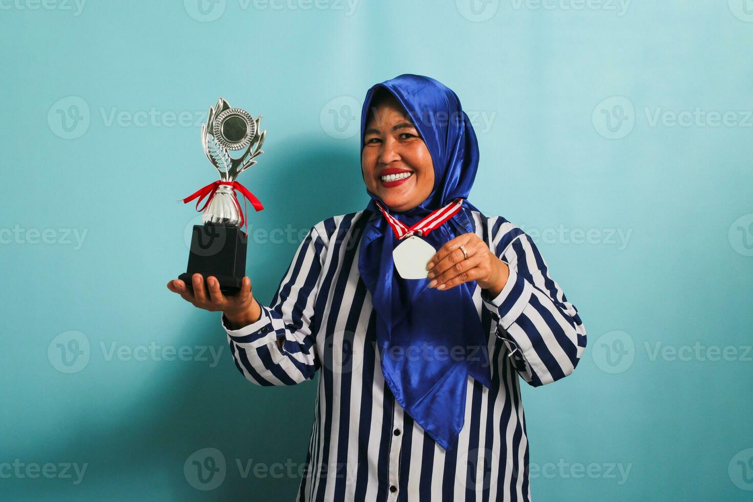 A happy middle-aged Asian businesswoman in a blue hijab and a striped shirt is showing an empty white medal while holding a silver trophy, celebrating her success, isolated on a blue background photo