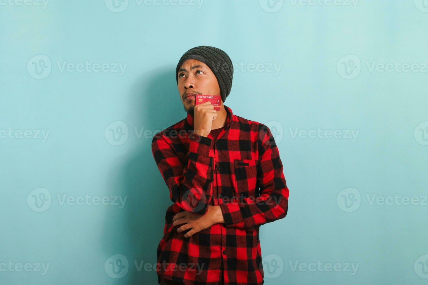 Pensive young Asian man is holding credit cards looking at copy space, isolated on blue background photo