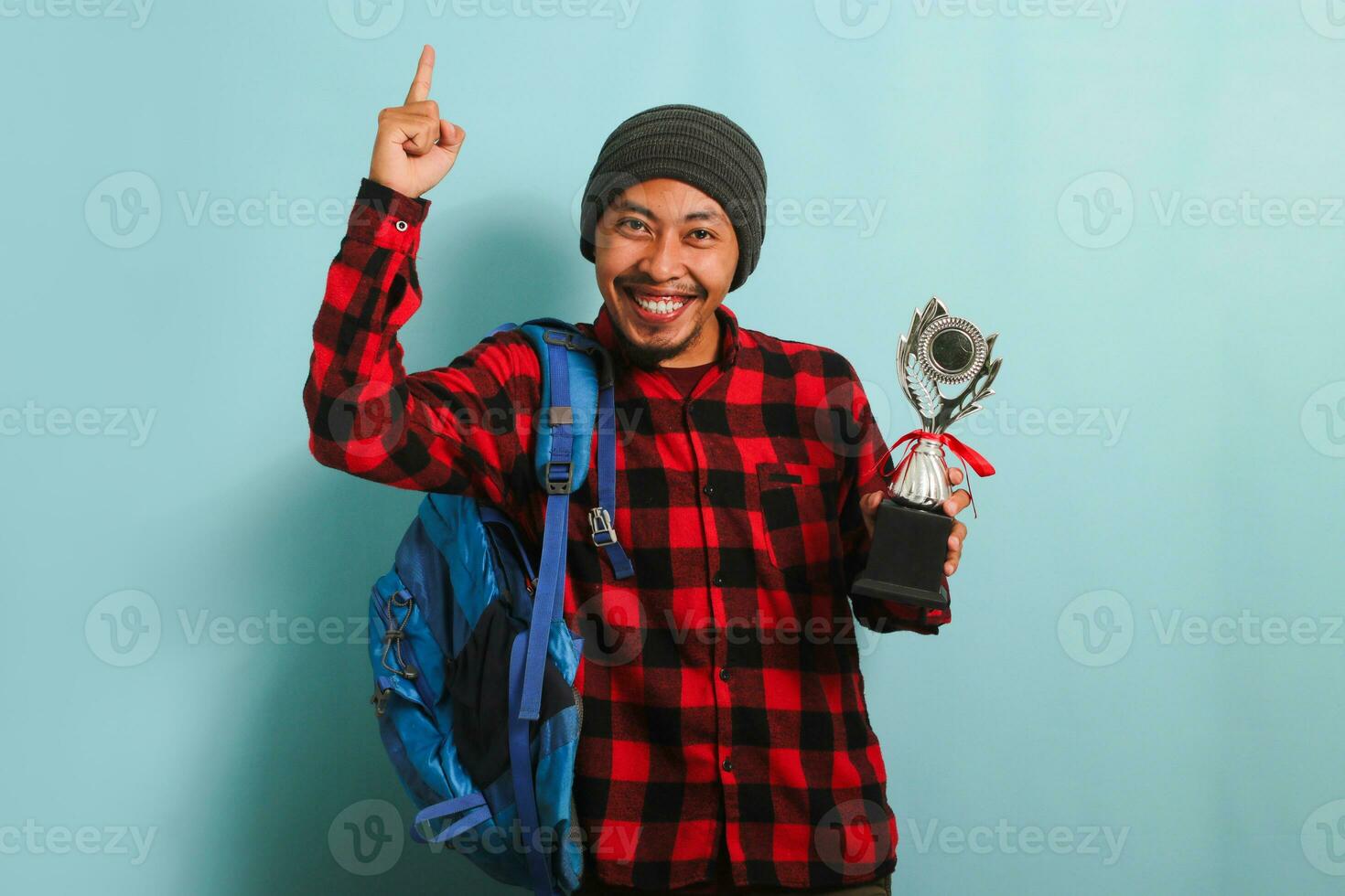 Excited Young Asian man student proudly pointing a trophy in his hand, isolated on blue background photo