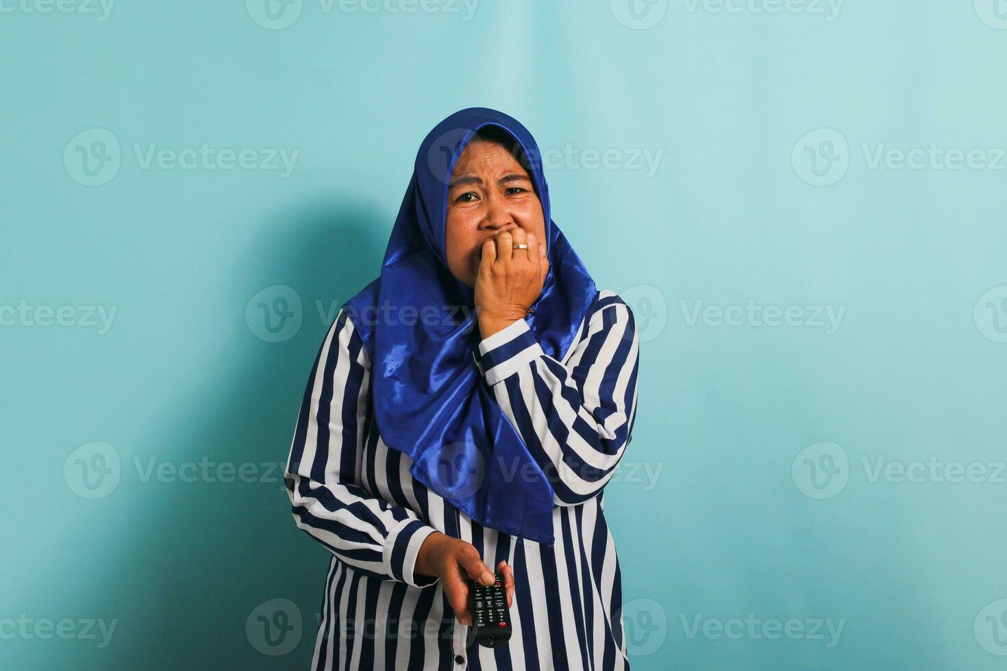 A worried middle-aged Asian woman in a blue hijab and a striped shirt is biting her fingers while holding a remote control and watching a horror movie on TV. She is isolated on a blue background photo