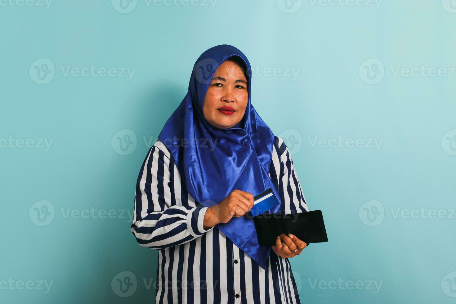 un contento de edad mediana asiático mujer en un azul hijab y un a rayas camisa toma fuera un crédito tarjeta desde su billetera a orden y pagar por adelantado para bienes en línea a un web almacenar, aislado en azul antecedentes foto