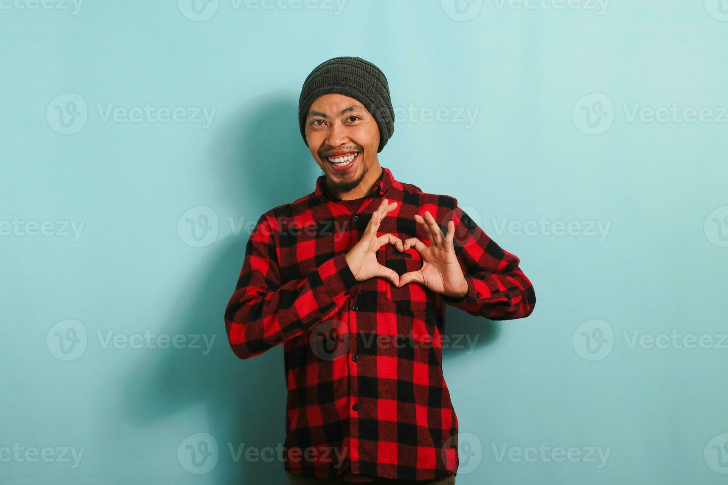 Smiling young Asian man shows a love gesture with his hand while standing against a blue background photo