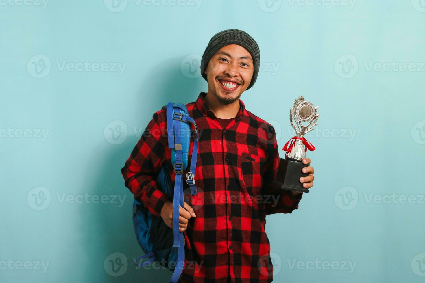 emocionado joven asiático hombre estudiante participación un trofeo, sonriente a el cámara, aislado en azul antecedentes foto