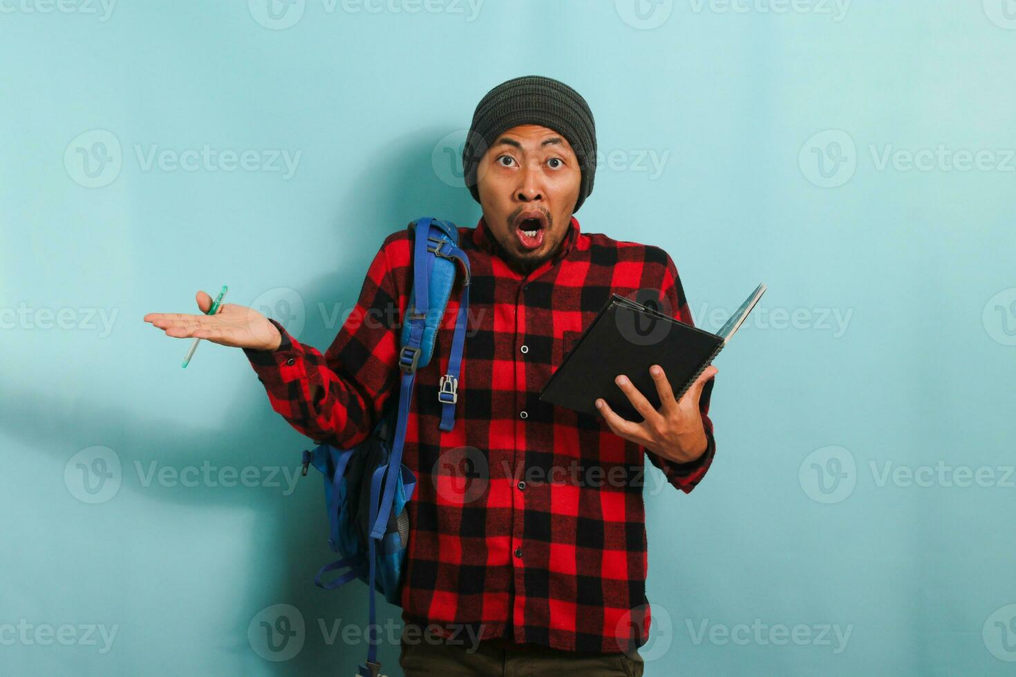 A shocked young Asian student with a beanie hat and a red plaid flannel shirt, wearing a backpack, raises his hands asking WHY to the camera while standing against a blue background photo