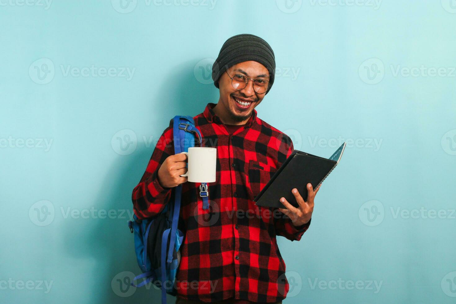 A young Asian male student with a beanie hat and a red plaid flannel shirt, wearing a rucksack and eyeglasses, takes a coffee break after lectures while standing against a blue background photo