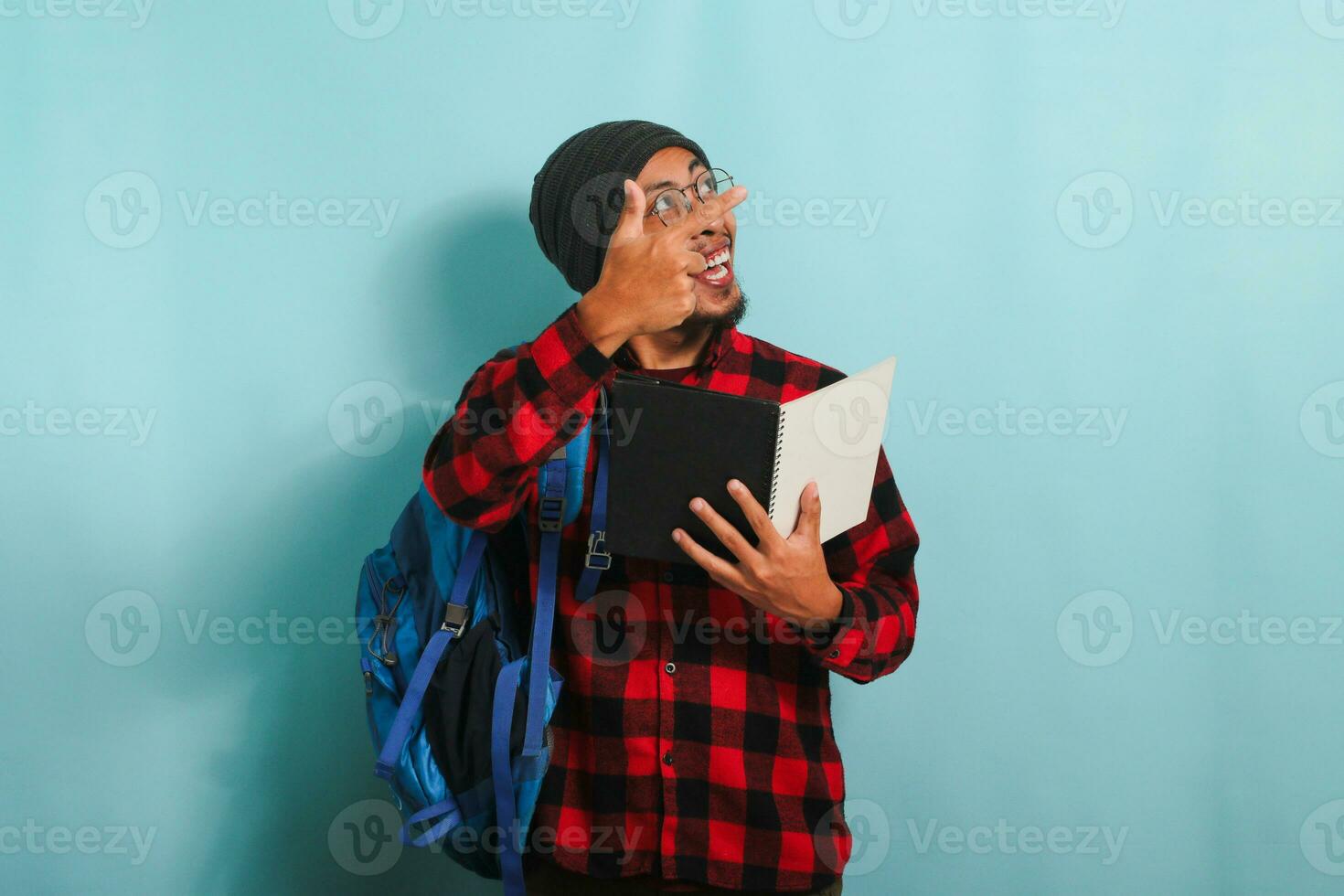 Excited young Asian student with a beanie hat and a red plaid flannel shirt, wearing a backpack and glasses, is holding books and pointing aside the copy space while standing against a blue background photo