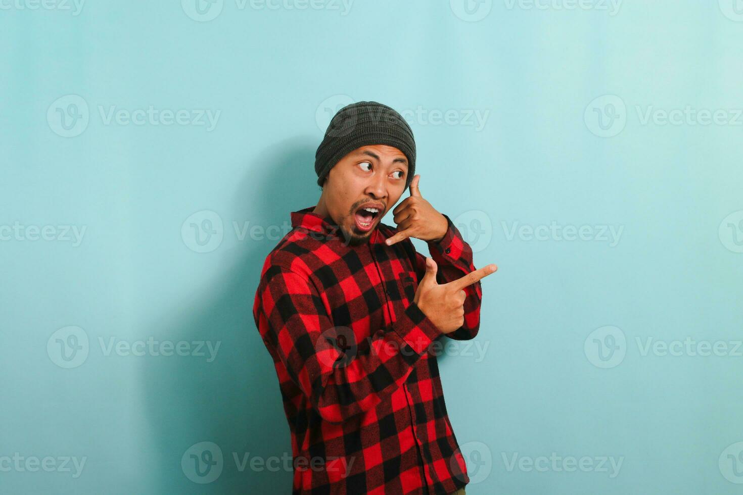 Excited young Asian man makes call me gesture, pointing to the right, isolated on a blue background photo