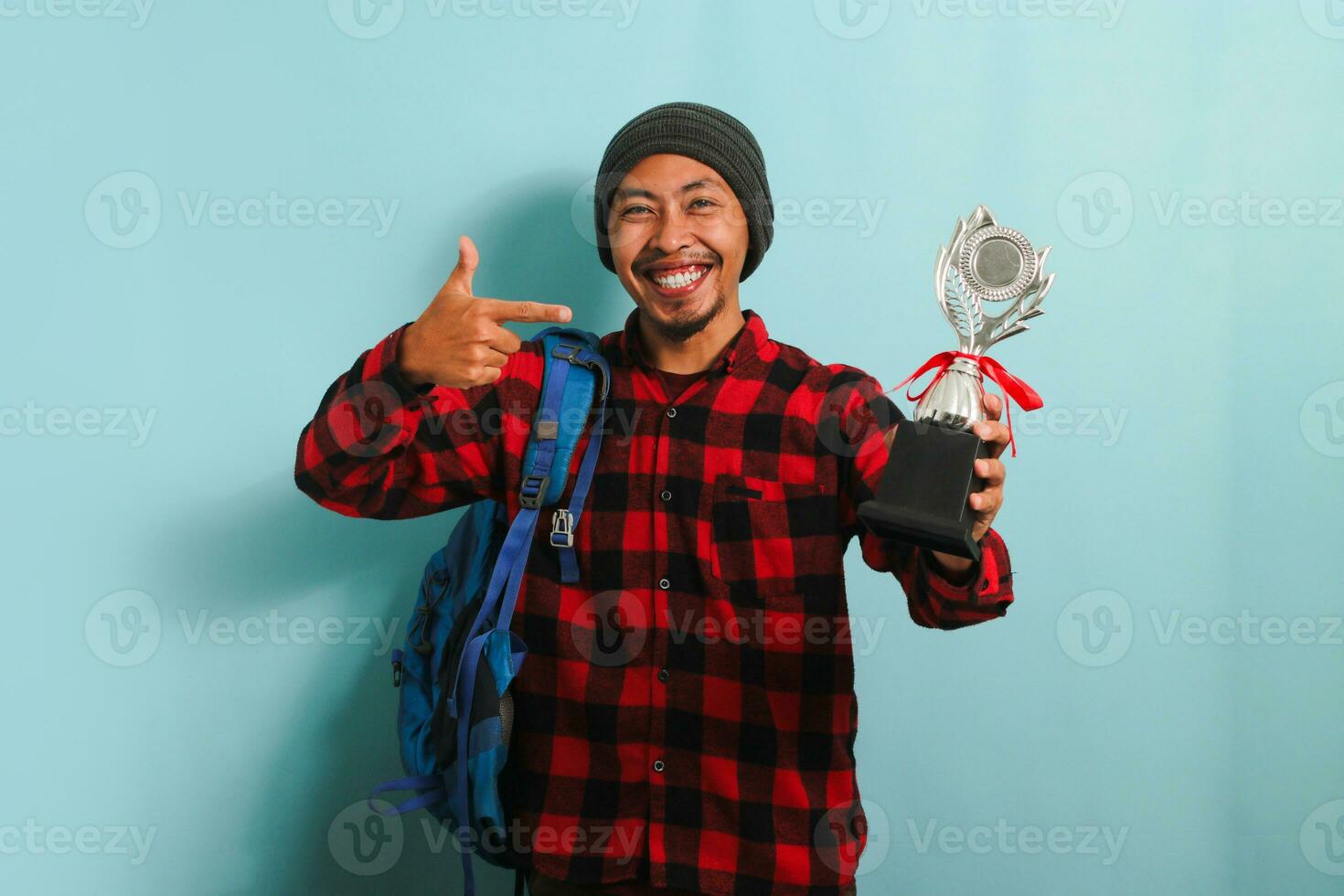 Excited Young Asian man student proudly pointing a trophy in his hand, isolated on blue background photo