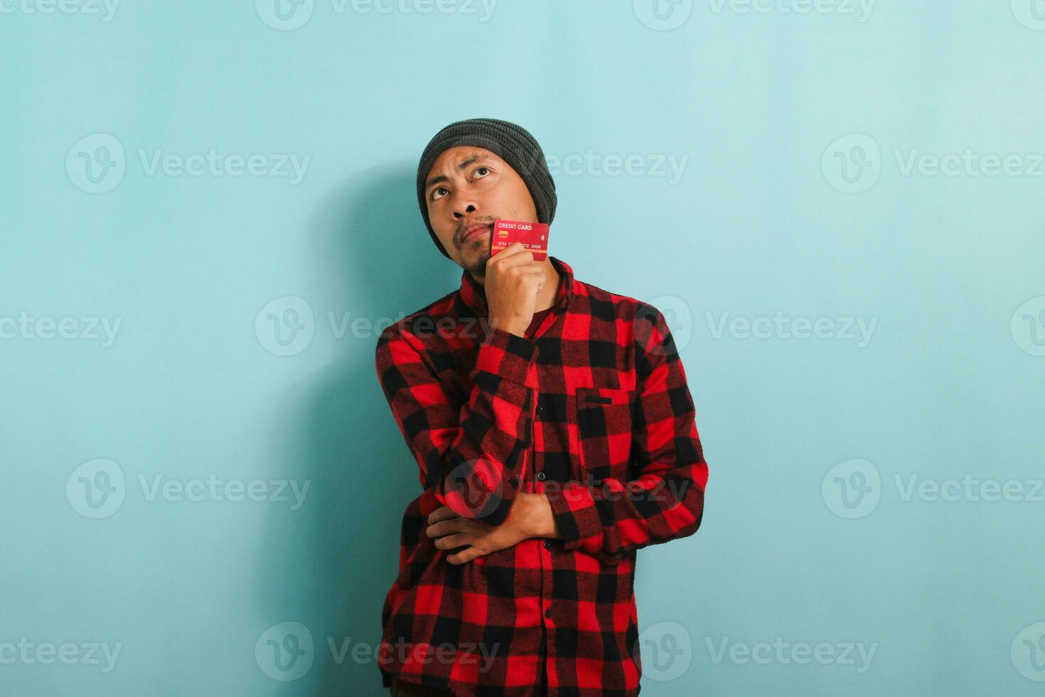 Pensive young Asian man is holding credit cards looking at copy space, isolated on blue background photo