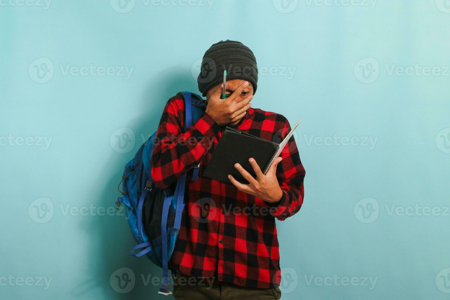 Angry young Asian student with a beanie hat and a red plaid flannel shirt, wearing a backpack, looks shocked after realizing he forgot to study before the exam while standing against a blue background photo