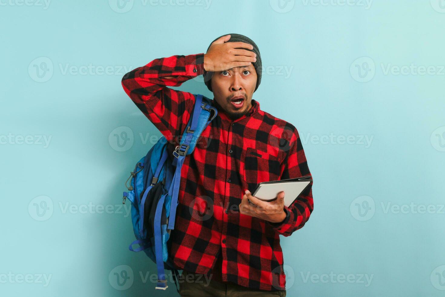 Surprised young Asian student appears unhappy with his hand on his head, isolated on blue background photo