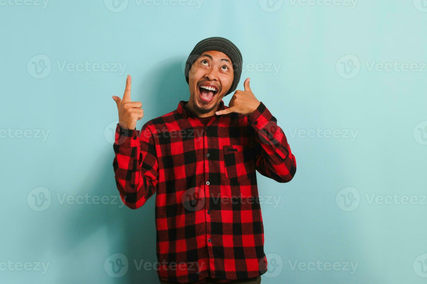 An excited young Asian man wearing a beanie hat and a red plaid flannel shirt makes a call me gesture, pointing up to the copy space, isolated on a blue background photo