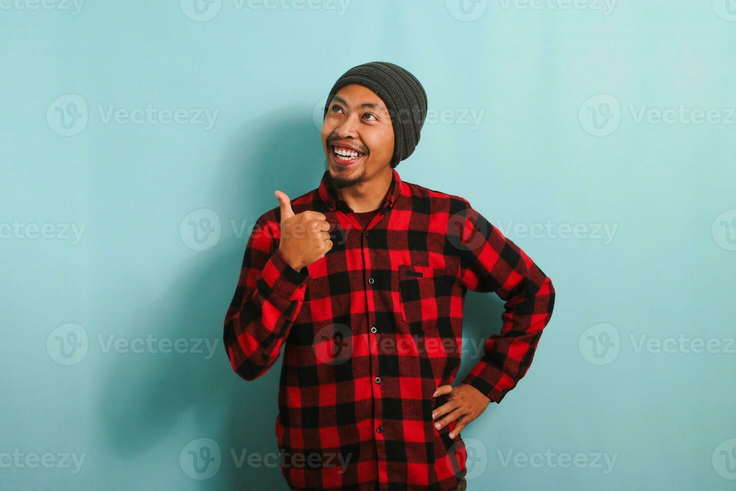 Confident young Asian man with a beanie hat and a red plaid flannel shirt is pointing his finger to the side empty copy space, while also giving a thumbs-up gesture, isolated on a blue background photo