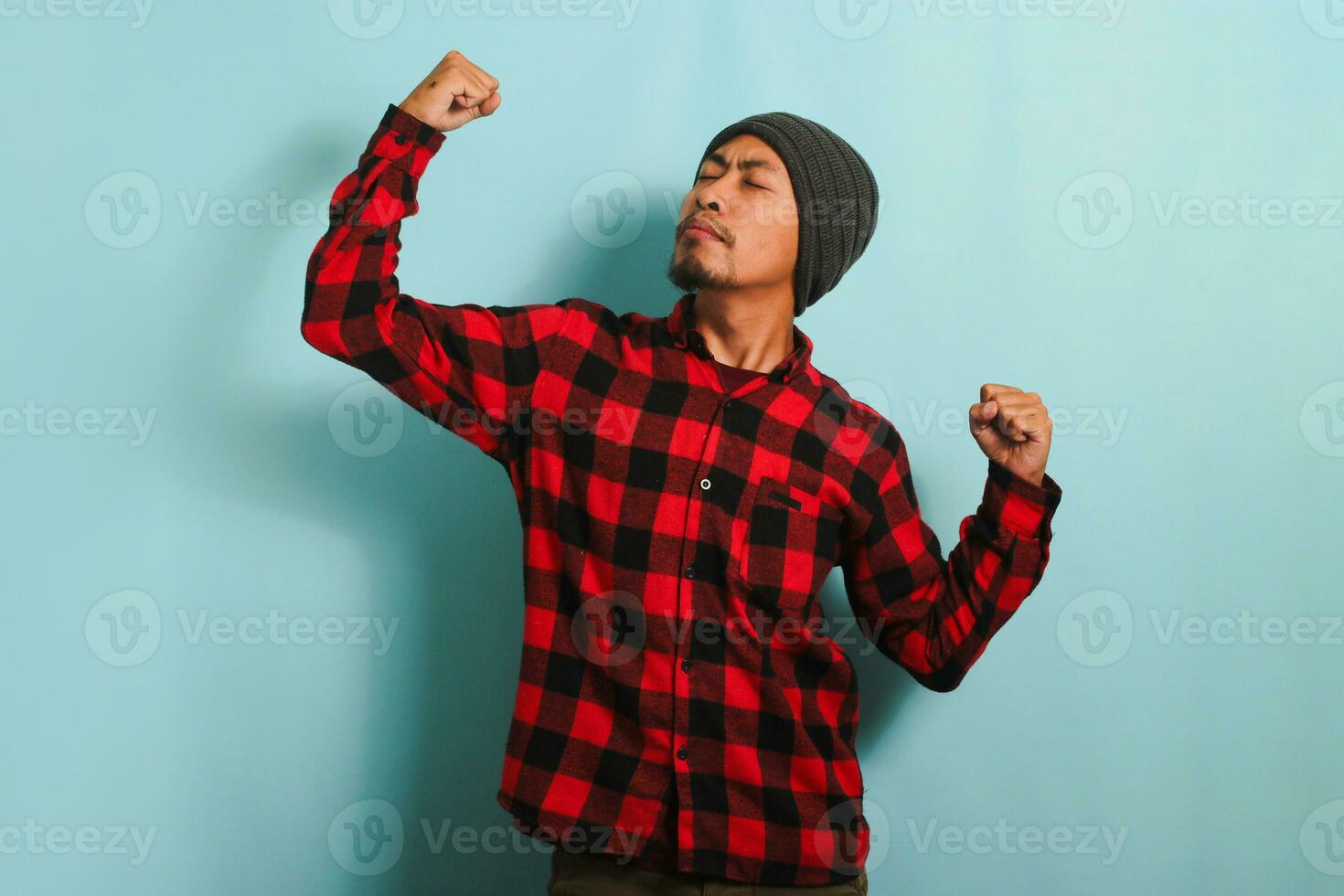 confidente joven asiático hombre con gorro sombrero y rojo tartán franela camisa haciendo un fuerte gesto, cerca su ojos, demostración fuerza por flexionando su brazos y músculos, aislado en un azul antecedentes foto