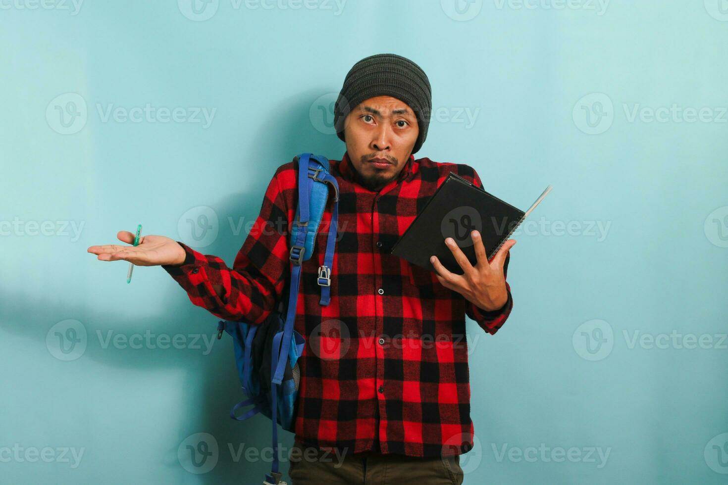 A shocked young Asian student with a beanie hat and a red plaid flannel shirt, wearing a backpack, raises his hands asking WHY to the camera while standing against a blue background photo