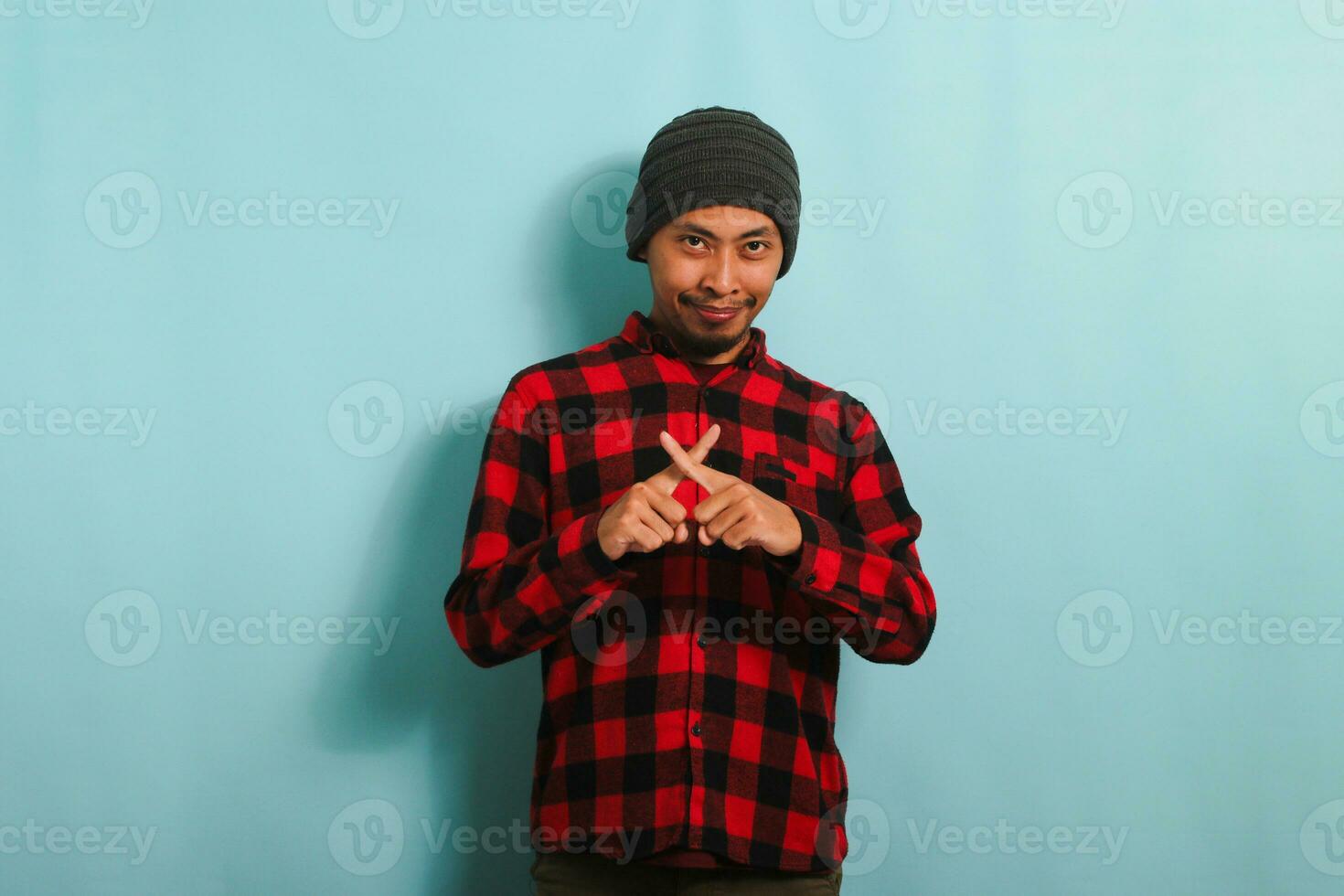 Serious young Asian man with beanie hat and red plaid flannel shirt showing a warning gesture, indicating to stop, decline, or forbid something, isolated on a blue background photo