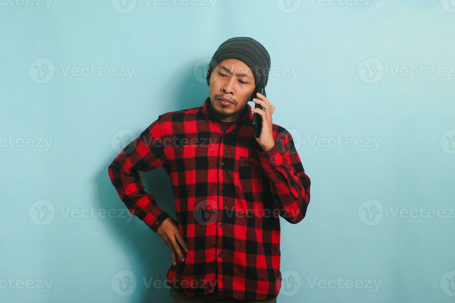 Serious Young Asian man with a beanie hat and a red plaid flannel shirt has a worried and unhappy look while talking on his mobile phone and receiving bad news, isolated on a blue background photo