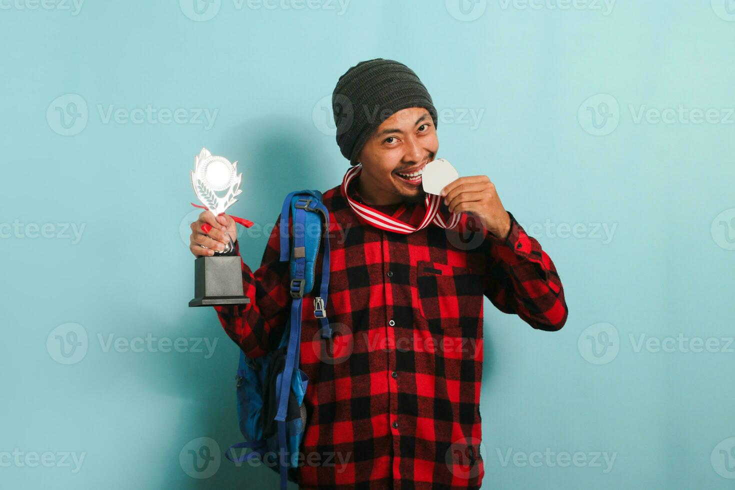 Happy young Asian man student wearing a backpack, beanie hat, and red plaid flannel shirt, biting a medal while holding a trophy, rejoicing in success and achievement, isolated on a blue background photo