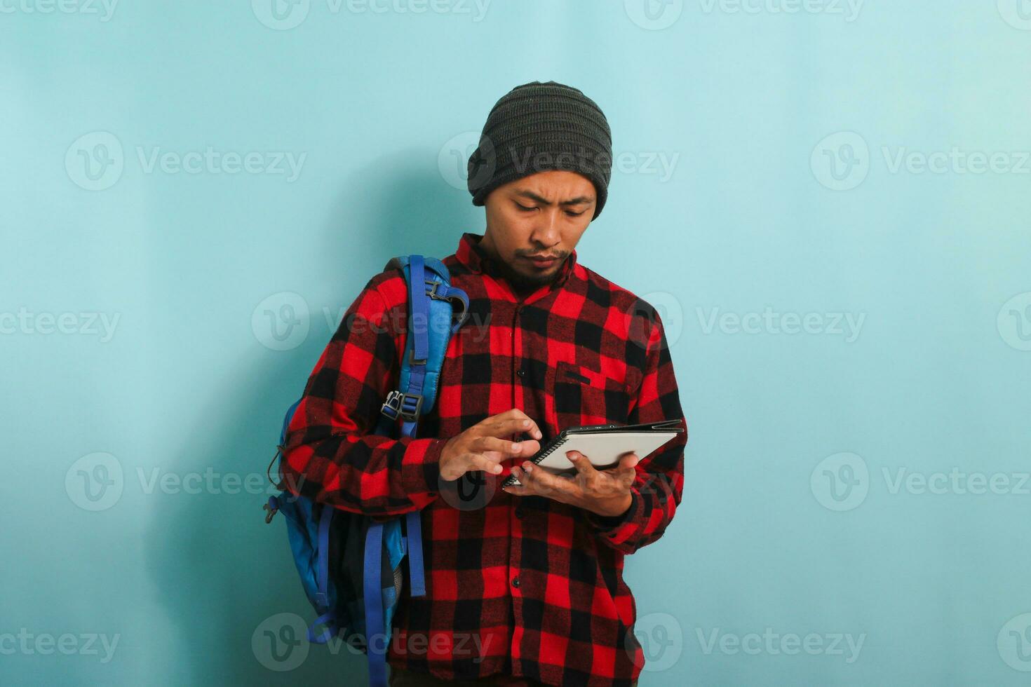 Pensive young Asian student with a beanie hat and a red plaid flannel shirt, wearing a backpack, is searching for ideas on the internet to finish his assignment isolated on blue background photo