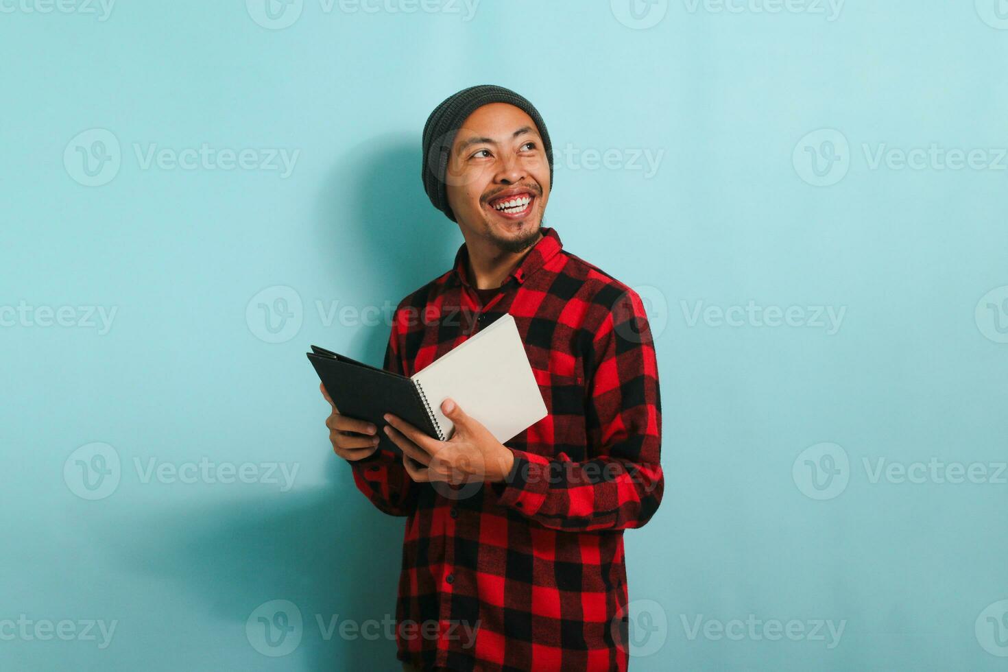 A smiling young Asian man with a beanie hat and a red plaid flannel shirt is holding a book while standing against a blue background photo