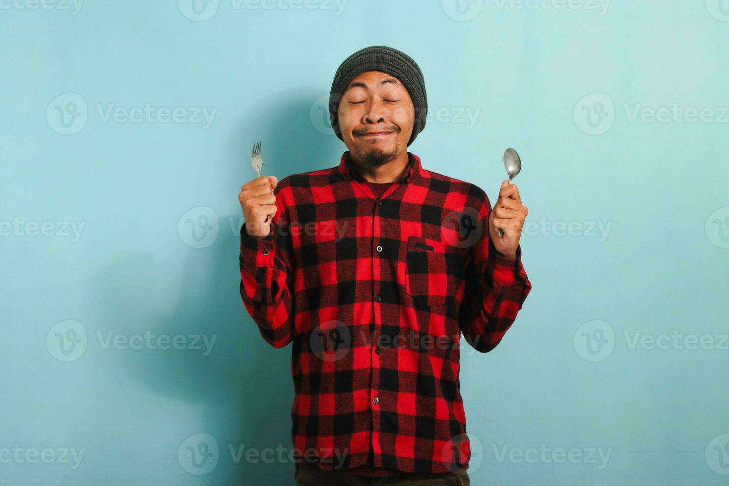 hambriento joven asiático hombre con gorro sombrero y rojo tartán franela camisa clausura su ojos, imaginando delicioso alimento, mientras participación un cuchara y tenedor, aislado en un azul antecedentes foto