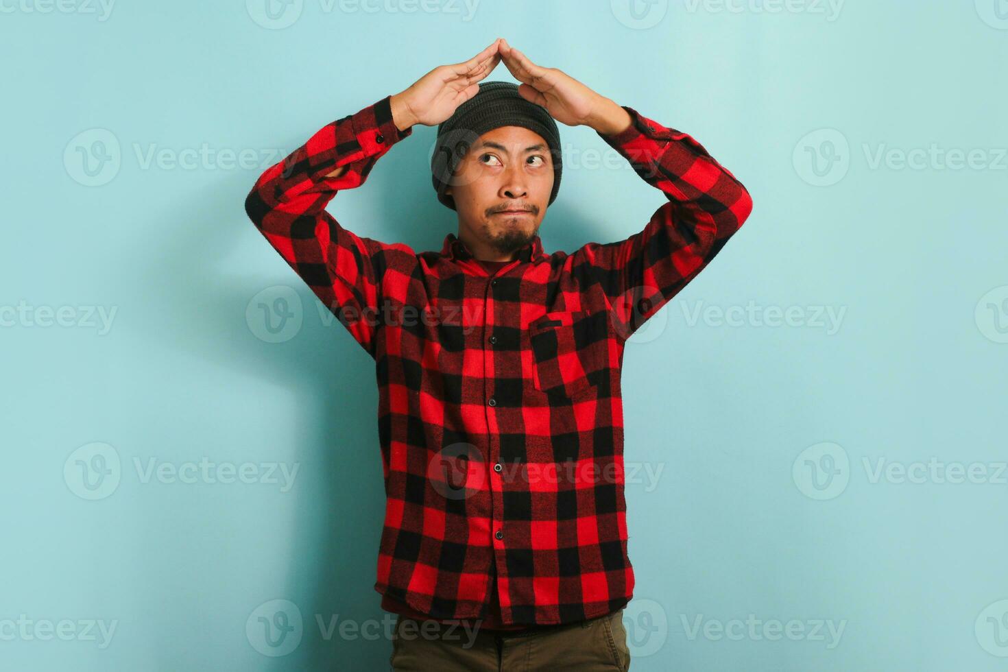 Pensive young millennial Asian man with a beanie hat and red plaid flannel shirt making a roof gesture above his head, thinking and planning to have his own home, isolated on a blue background. photo