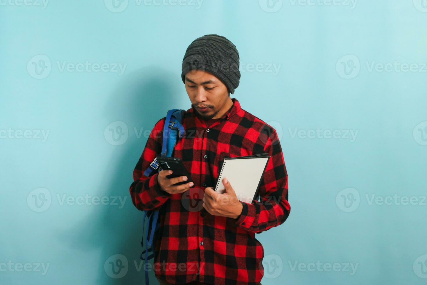 Pensive young Asian student with a beanie hat and a red plaid flannel shirt, wearing a backpack, is searching for ideas on the internet to finish his assignment isolated on blue background photo