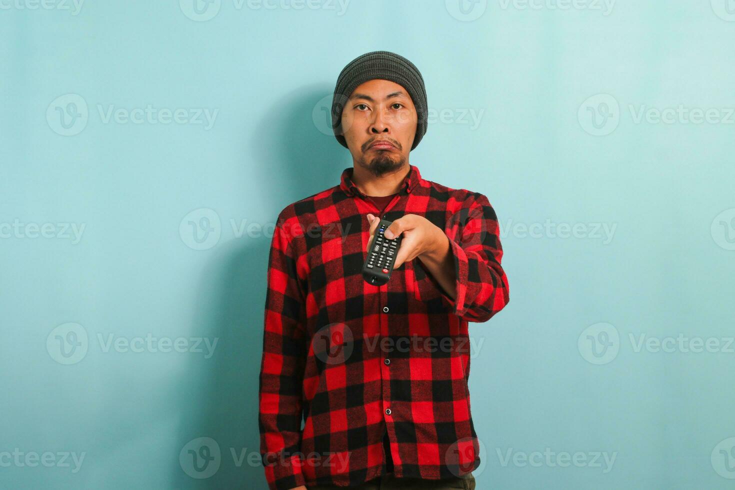 An annoyed young Asian man with a beanie hat and red plaid flannel shirt is switching TV channels using the TV remote, feeling upset and bored, isolated on a blue background. photo