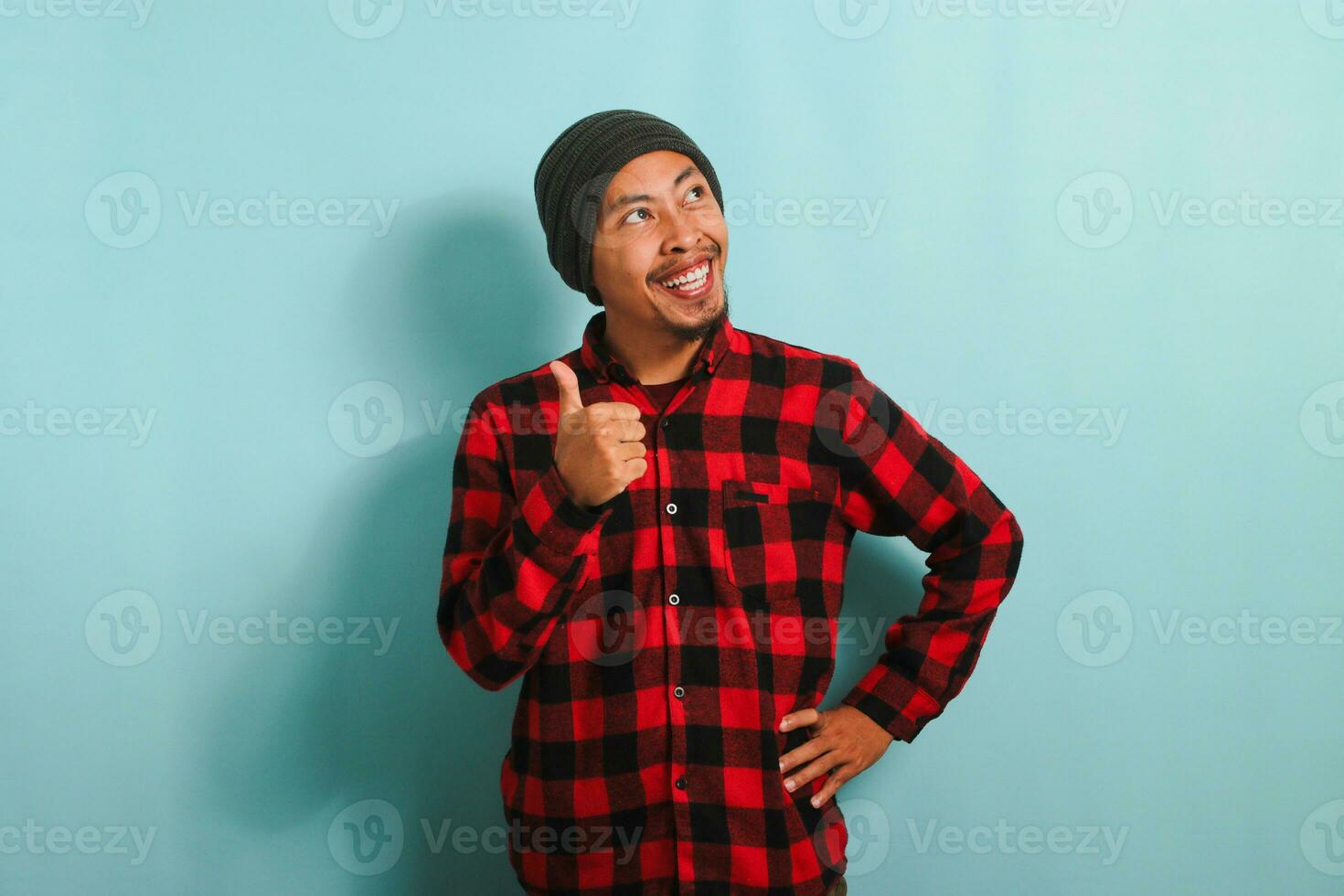 A confident young Asian man with a beanie hat and a red plaid flannel shirt is giving a thumbs-up while looking at right copy space, expressing a positive review. He is isolated on a blue background photo